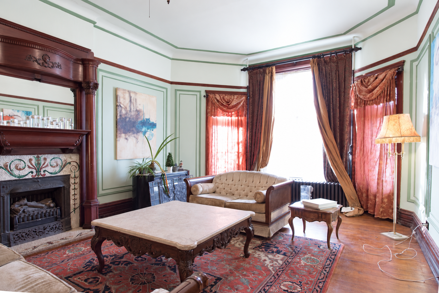 The house’s Victorian parlor with carved wood fireplace, sage trim, and seating beside tall windows